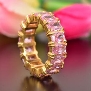 May include: A gold ring with a row of pink square-shaped gemstones. The ring is set on a dark brown surface.
