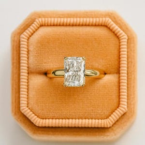 May include: A gold ring featuring a rectangular-cut diamond, presented in a peach velvet box. The diamond sparkles, catching the light. The ring box has a textured, ribbed border, adding to its luxurious appearance.