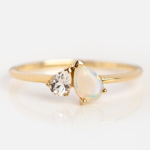 May include: A gold ring with two teardrop-shaped stones. The larger stone is a white opal, and the smaller stone is a clear quartz.