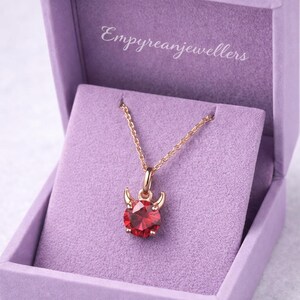 May include: A gold-colored necklace with a red gemstone pendant shaped like a devil. The pendant has small horns and is set in a gold-colored metal. The necklace is displayed in a purple jewelry box. The text "Empyreanjejewellers" is visible.