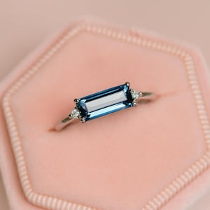 Puede incluir: Un anillo de plata con una piedra preciosa rectangular de color azul oscuro como pieza central. Pequeñas piedras de acento claras flanquean la piedra principal a ambos lados. El anillo se presenta sobre un fondo rosa suave, lo que sugiere una exhibición de joyas.