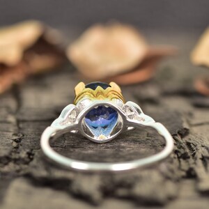 Op de afbeelding: Een zilveren ring met een grote, ovaalvormige blauwe edelsteen. De ring heeft een goudkleurige zetting rond de edelsteen en decoratieve zilveren accenten op de band. De ring staat tegen een wazige achtergrond.