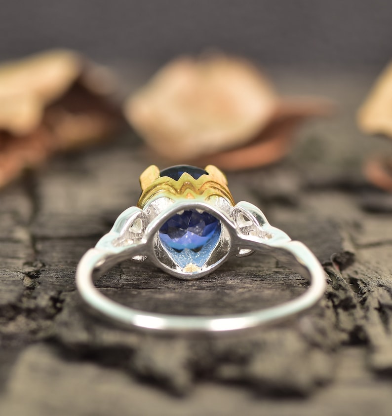 Op de afbeelding: Een zilveren ring met een grote, ovaalvormige blauwe edelsteen. De ring heeft een goudkleurige zetting rond de edelsteen en decoratieve zilveren accenten op de band. De ring staat tegen een wazige achtergrond.