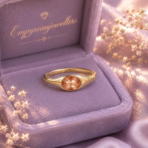 May include: A gold ring with an oval-shaped peach-colored gemstone set in a smooth band. The ring is displayed inside a lavender velvet jewelry box with the text Empyreanjewellers printed in gold on the inside lid, accompanied by small white flowers.
