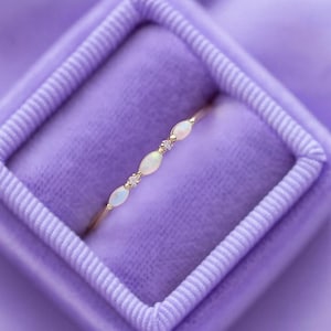 Könnte beinhalten: Ein zarter goldener Ring mit ovalen Opalen und kleinen Akzentsteinen. Der Ring wird in einer lila Samtbox präsentiert, die sein elegantes Design hervorhebt. Die Opale sind milchig weiß. Das Design des Rings ist elegant und raffiniert.