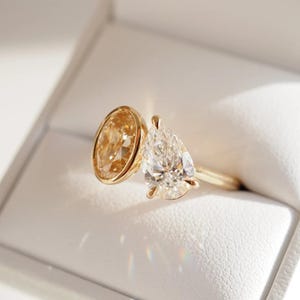 May include: A gold ring with two gemstones: a clear, pear-shaped diamond and an oval-cut yellow diamond. The ring is set in a gold band and displayed in a white jewellery box, highlighting the ring's sparkle and design.