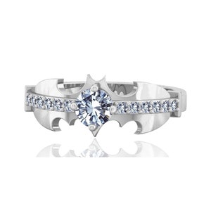 Peut inclure: Une bague en argent avec une grande pierre précieuse ronde et transparente au centre. La bague est en forme de symbole de chauve-souris avec une rangée de petites pierres précieuses autour de la bande.