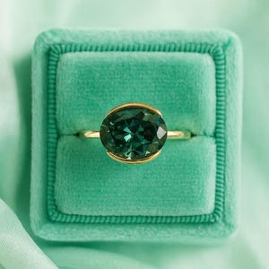 Puede incluir: Un anillo de oro con una gran piedra preciosa ovalada de color verde azulado. El anillo está engastado en un bisel de oro y se encuentra en un joyero de terciopelo verde azulado. El fondo es una tela suave y verde azulado claro.