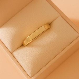 May include: A gold ring with a rectangular design, presented in a beige jewellery box. The ring has a smooth, polished surface and a slightly curved shape. The box has a soft, leather-like texture.