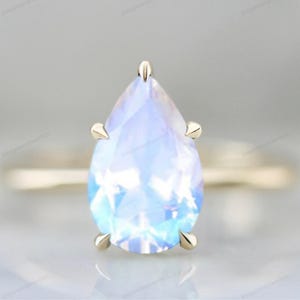 May include: A pear-shaped moonstone engagement ring set in a gold band. The ring features a prong setting and a clear, iridescent stone.