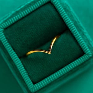 Könnte beinhalten: Ein goldfarbener, V-förmiger Ring liegt in einer dunkelgrünen Samt-Ringbox. Der Ring hat ein schlichtes, elegantes Design. Die Box hat eine quadratische Form mit einem strukturierten, gerippten Rand.