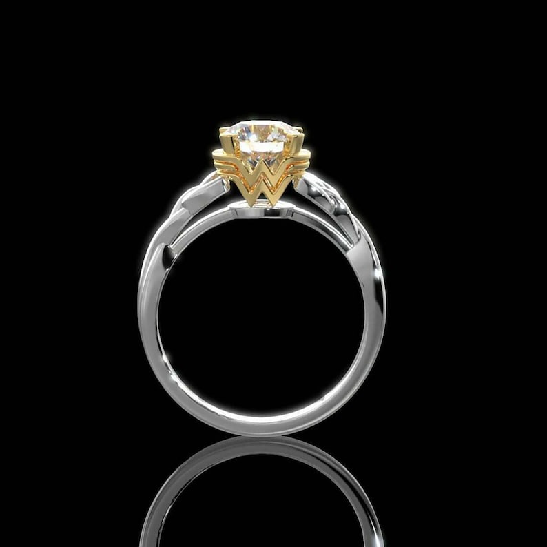 Op de afbeelding: Een zilveren ring met een gouden accent met een grote, ronde, heldere edelsteen. Het gouden accent heeft de vorm van een gestileerde "W". De ring staat tegen een zwarte achtergrond, met een weerspiegeling eronder zichtbaar.