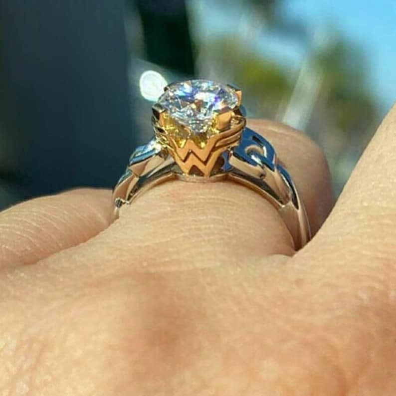 Op de afbeelding: Een close-up van een diamanten ring. De ring heeft een grote, ronde, heldere diamant gezet in een gouden en zilveren band. De band heeft een ingewikkeld ontwerp met gouden accenten rond de diamantzetting.