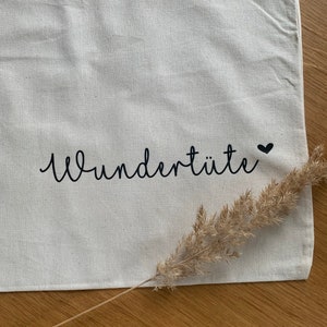 Könnte beinhalten: Eine weiße Stofftasche mit dem Text "wunderhüte" in schwarzer, kursiv geschriebener Schrift und einem kleinen schwarzen Herz. Der Text ist etwa 19 cm breit.