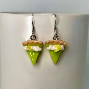 May include: A pair of lime green key lime pie earrings with a white meringue topping and a lime slice on top. The earrings are made of polymer clay and have silver hooks.