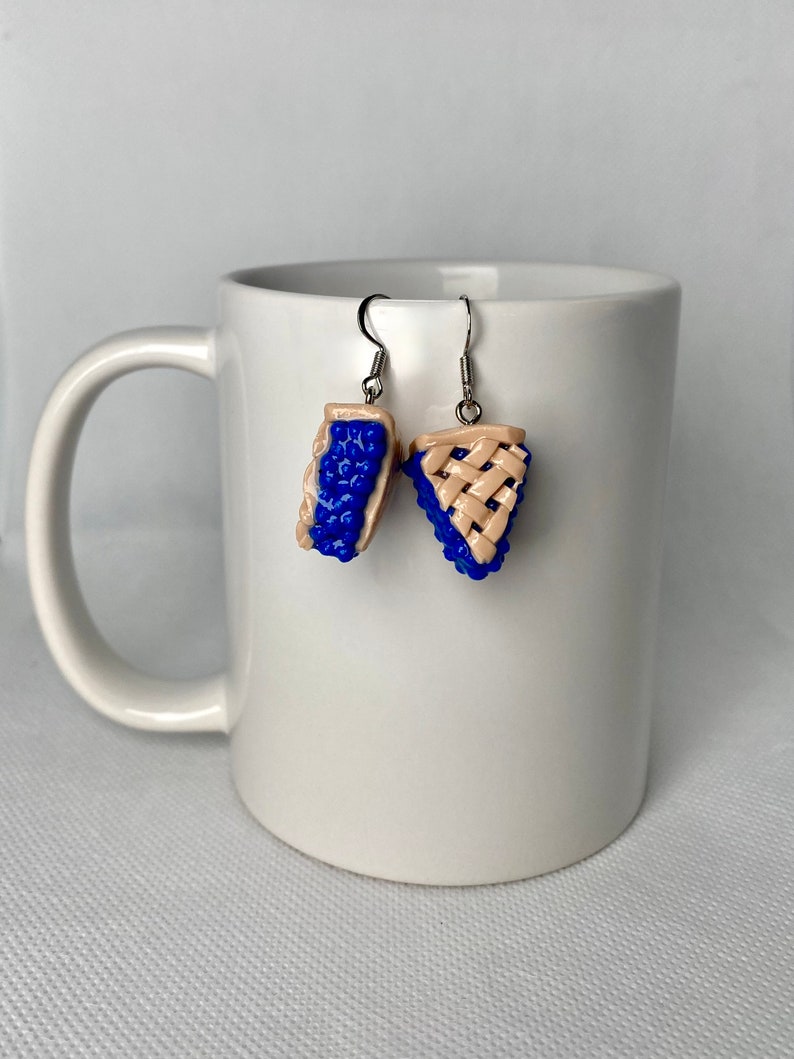 May include: A pair of dangle earrings featuring a blueberry pie design. The earrings are made of polymer clay and have a silver tone finish.
