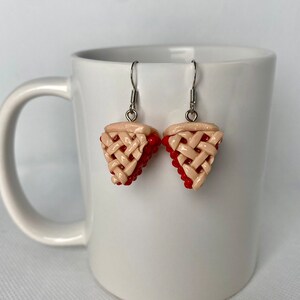 May include: A pair of dangle earrings featuring a red and beige cherry pie design. The earrings are made of polymer clay and have silver hooks.