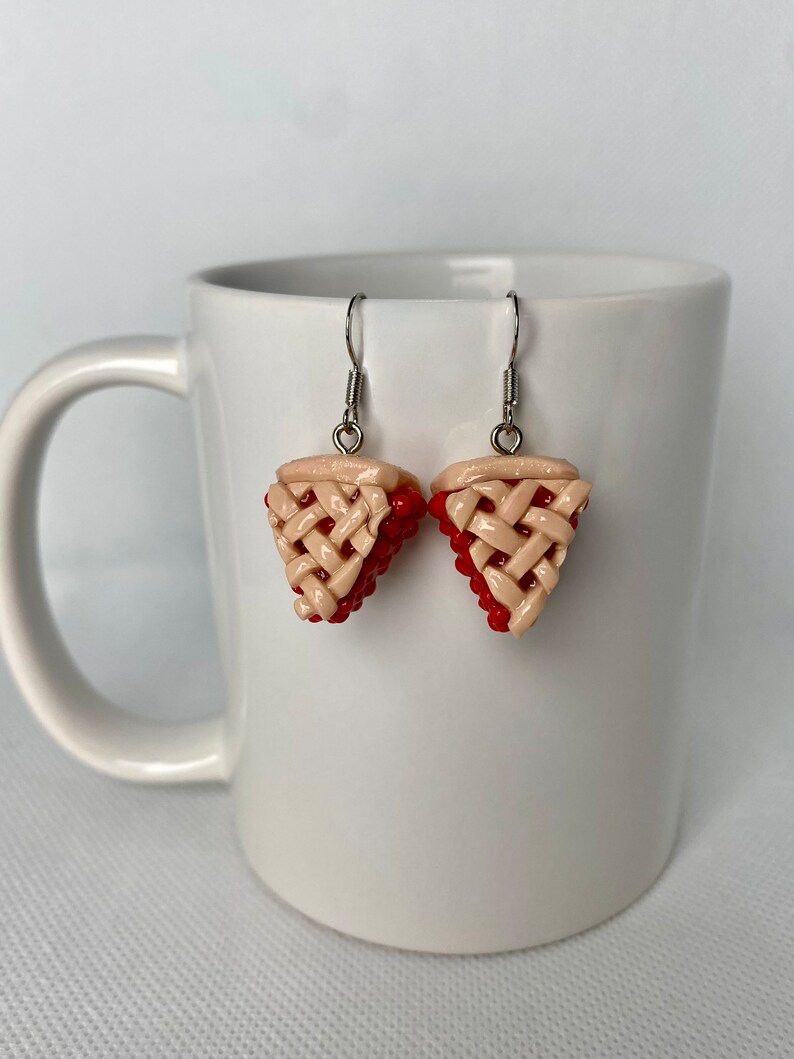 May include: A pair of dangle earrings featuring a red and beige cherry pie design. The earrings are made of polymer clay and have silver hooks.