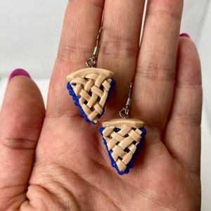May include: A pair of blue and beige dangle earrings shaped like blueberry pie slices. The earrings have a woven crust design and are made of polymer clay.
