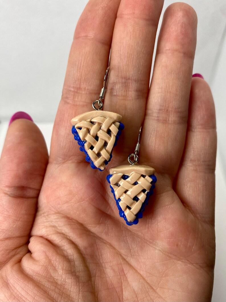 May include: A pair of blue and beige dangle earrings shaped like blueberry pie slices. The earrings have a woven crust design and are made of polymer clay.