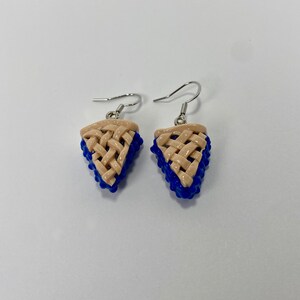 May include: A pair of silver dangle earrings featuring a blue and beige pie slice design. The pie crust is woven and the filling is blue.