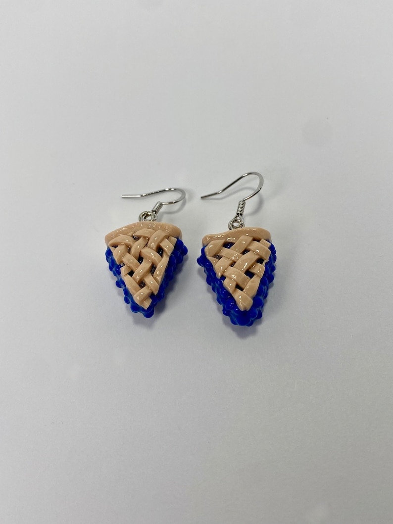 May include: A pair of silver dangle earrings featuring a blue and beige pie slice design. The pie crust is woven and the filling is blue.