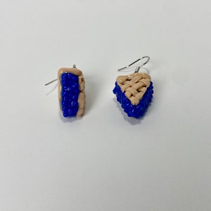 May include: A pair of blue and beige dangle earrings shaped like blueberry pie slices.