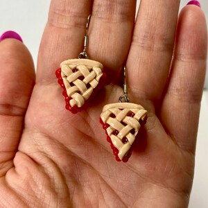 May include: A pair of dangle earrings featuring a cherry pie slice design. The earrings are made of polymer clay and have a realistic look. The pie crust is a light beige color and the cherries are a bright red.
