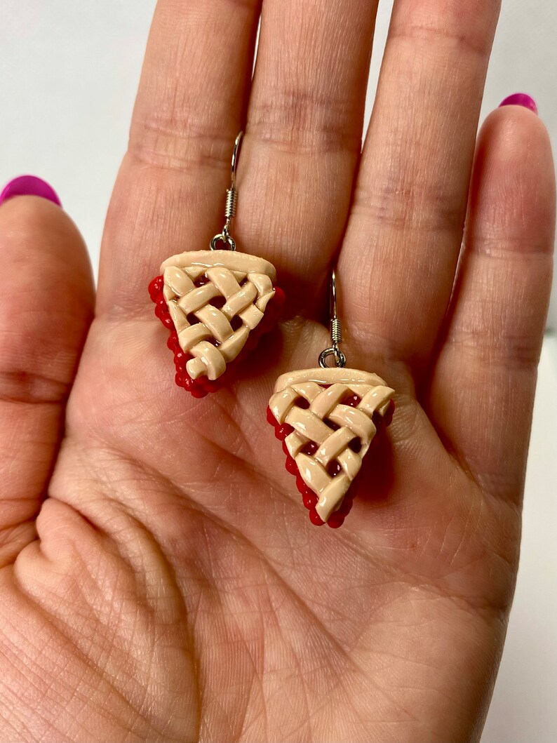 May include: A pair of dangle earrings featuring a cherry pie slice design. The earrings are made of polymer clay and have a realistic look. The pie crust is a light beige color and the cherries are a bright red.