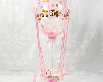 May include: A decorative hot air balloon ornament. The clear balloon is filled with pink and gold confetti and has "Happy Birthday" written on it. It is attached to a white wicker basket with pink ribbons and a bow.