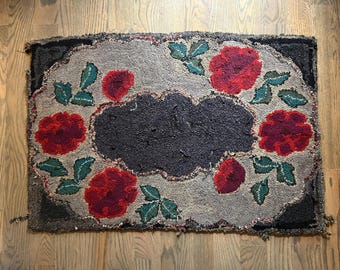 Puede incluir: Alfombra hecha a mano con un diseño floral. La alfombra presenta un centro y un borde gris oscuro, con un fondo gris claro. Rosas rojas y hojas verdes están dispuestas alrededor del centro. La alfombra es rectangular y descansa sobre un suelo de madera.