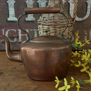 May include: A vintage copper teapot with a brass lid and handle. The teapot has a rounded body and a spout on the side. The teapot is sitting on a wooden table with a wicker bottle and yellow flowers in the background.