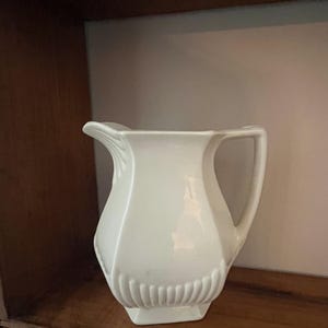 Op de afbeelding: Een witte keramische kan met een handvat en een tuit. De kan heeft een geometrisch ontwerp met verticale ribbels nabij de basis. De kan staat op een houten plank. De kan wordt waarschijnlijk gebruikt voor het serveren van dranken.
