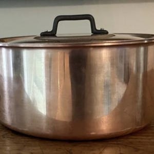 Puede incluir: Una olla de cobre con tapa y dos asas laterales. La olla tiene una superficie brillante y reflectante y asas de color oscuro. La tapa también tiene un asa oscura. La olla está sobre una superficie de madera.