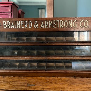 Può includere: Mobile portaoggetti vintage in legno con tre cassetti. Il mobile presenta la scritta "BRAINERD & ARMSTRONG CO." in lettere dorate. Ogni cassetto ha più scomparti e un frontale in vetro. Il mobile è in legno scuro.