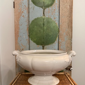 May include: A white ceramic tureen featuring a wide, rounded bowl, a pedestal base, and two decorative scrolled handles on the sides. The exterior surface of the bowl displays subtle vertical paneling and embossed leaf details near the handles.
