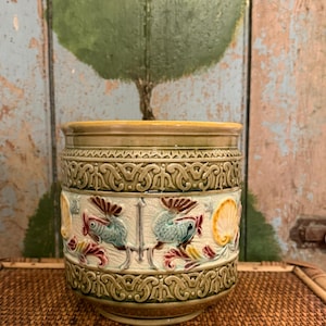 May include: A cylindrical ceramic planter featuring a raised relief design of stylized blue fish and yellow seashells against a cream-colored crackle glaze background, framed by intricate olive green geometric borders at the top and bottom.