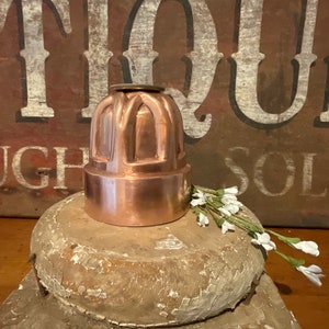 May include: A copper-colored, antique jelly mold with a decorative, arched design. The mold is sitting on a wooden surface.