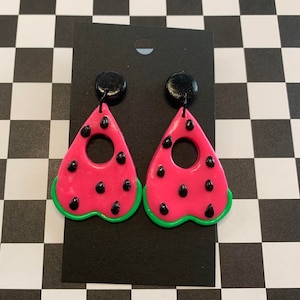 May include: A pair of pink watermelon-shaped earrings with black seeds and green edges. The earrings are in the shape of a heart with a circle cut out of the center.