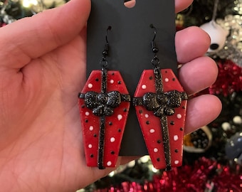 May include: Red coffin-shaped earrings with white polka dots and a black bow. The earrings have black ear wires and are displayed on a black card. The background includes a Christmas tree with red and black ornaments.