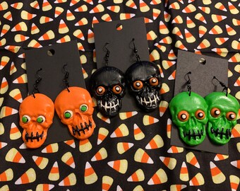 May include: Three pairs of Halloween-themed skull earrings. One pair is black, one pair is orange, and one pair is green. Each pair has large, round eyes and a stitched mouth.