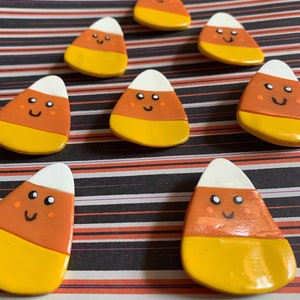 Könnte beinhalten: Sieben handgemachte Zuckermais-Ornamente, die in Weiß, Orange und Gelb bemalt sind. Jeder Zuckermais hat ein lächelndes Gesicht mit schwarzen Augen und einem schwarzen Mund.