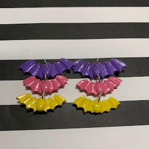 May include: A pair of dangle earrings featuring three layered bats. The top bat is purple, the middle bat is pink, and the bottom bat is yellow.