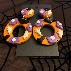 May include: A pair of orange Halloween-themed earrings with purple, white, and black accents. The earrings feature ghosts, bats, and flowers.