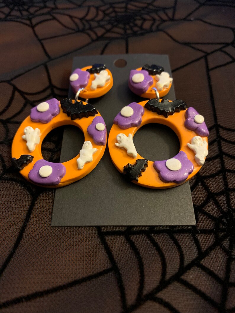 May include: A pair of orange Halloween-themed earrings with purple, white, and black accents. The earrings feature ghosts, bats, and flowers.