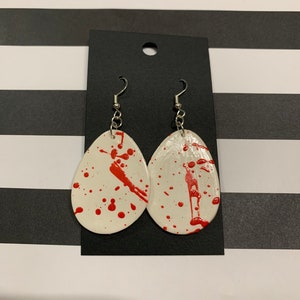 May include: A pair of white teardrop-shaped earrings with red paint splatters. The earrings have silver hooks.