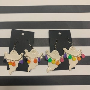 May include: White ghost-shaped earrings with smiling faces and colourful candy corn accents. Each earring has a silver chain holding the candy corn. The earrings are displayed on black earring cards against a black and white striped background.