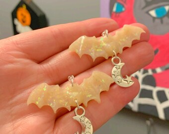 May include: A pair of iridescent white bat earrings with silver crescent moon charms. Each charm has five small stars.