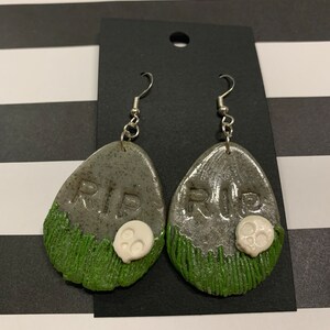 Puede incluir: Un par de pendientes de plata con un diseño de lápida gris y verde. Las lápidas tienen las palabras "RIP" y una calavera blanca en la parte inferior.