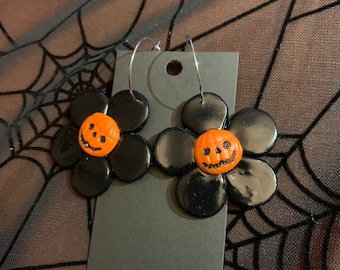 May include: A pair of black flower-shaped earrings with orange pumpkin faces. The earrings are hanging from silver hoops.